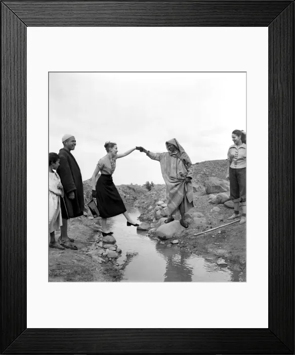 Framed Print : Man in arabic dress lends his hand to help French actresses across a stream as he guides Framed Print : Man in arabic dress lends his hand to help French actresses across a stream as he guides