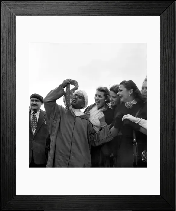 Framed Print : Local man of Morocco holding up a large snake near his mouth to the amusement of a group Framed Print : Local man of Morocco holding up a large snake near his mouth to the amusement of a group