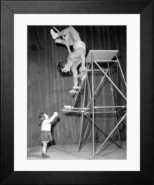 Framed Print : Children Acrobat. A young girl holding a cup looks up at a man walking down steps on his