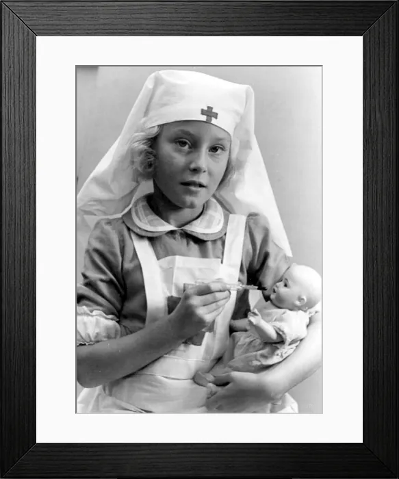 Framed Print : Young girl dressed in nurses outfit feeding her baby doll Circa 1945 P044506