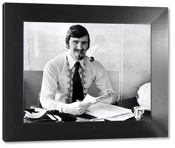 Jimmy Hill sitting at his desk smoking a pipe, dressed in stylish 1970s clothing