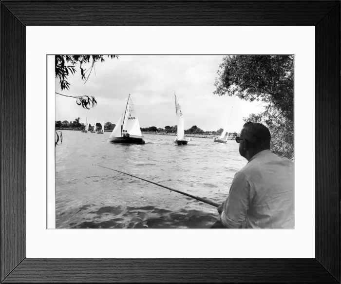 Framed Print : Angling. Under the cool shade of trees, a fisherman looks on at yachts as they cut Framed Print : Angling. Under the cool shade of trees, a fisherman looks on at yachts as they cut