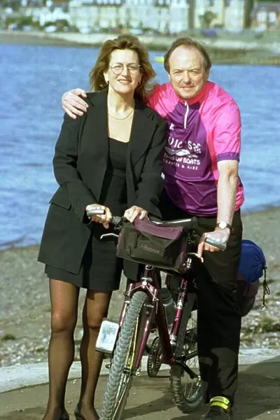 Barbara Dickson wearing glasses and a black suit poses with James Bolam on a bicycle in