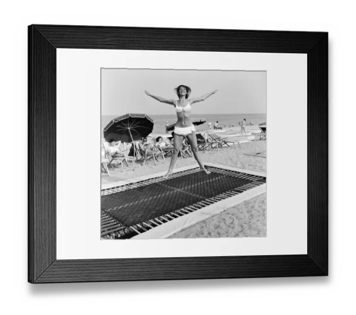 Framed Print : A model wearing bikini swimwear bounces on a trampoline on a beach in the French Riviera