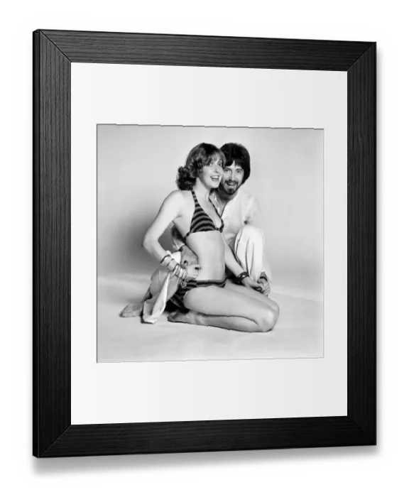 Framed Print : Couple wearing 1970s fashion. The woman wearing a matched striped bikini with the man Framed Print : Couple wearing 1970s fashion. The woman wearing a matched striped bikini with the man