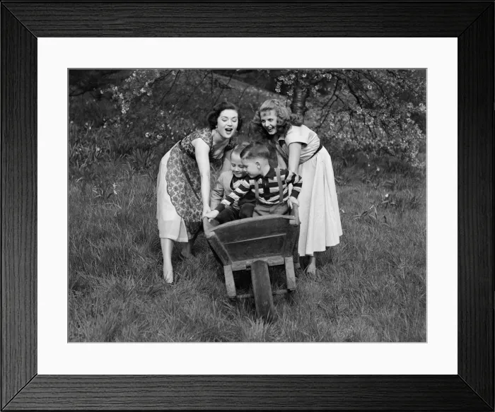 Framed Print : Two girls rolling two small boys down the hill in a wheelbarrow on a pleasant Spring day
