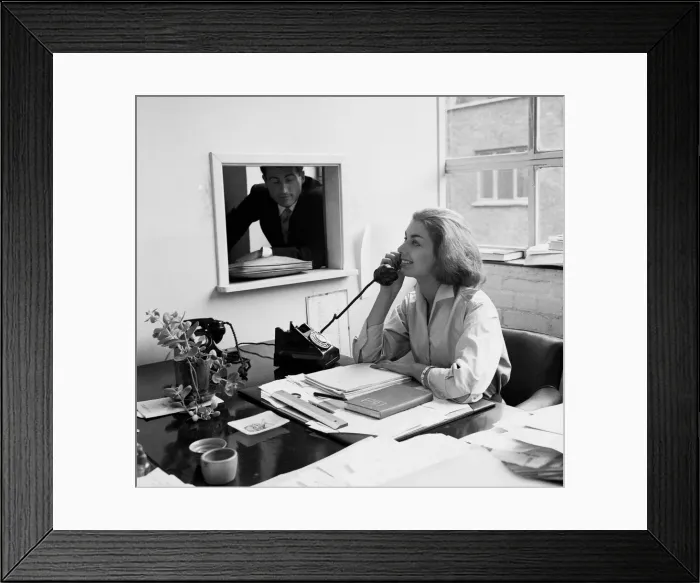 Framed Print : A woman making a phone call her office desk as a colleague looks through the window Framed Print : A woman making a phone call her office desk as a colleague looks through the window