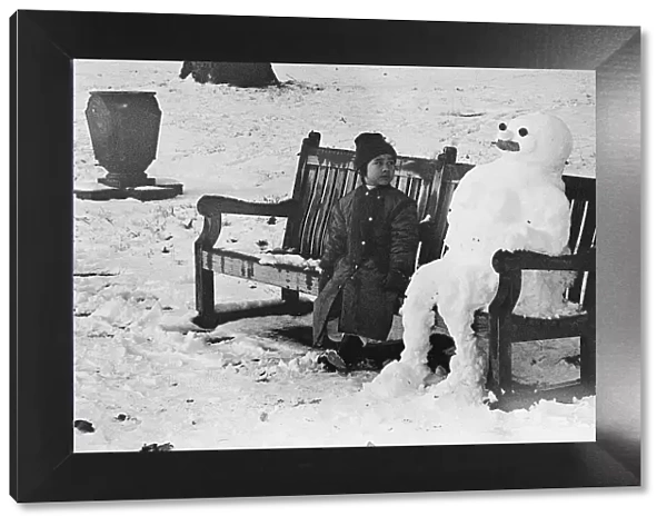 A little girls is captured sitting next to a Snow Man on a park bench