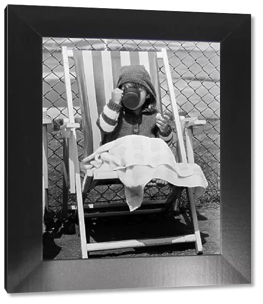 Young child swigs from a mug of juice, whilst sitting on a deck chair