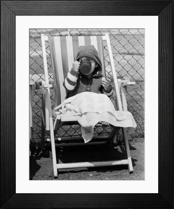 Framed Print : Young child swigs from a mug of juice, whilst sitting on a deck chair Framed Print : Young child swigs from a mug of juice, whilst sitting on a deck chair