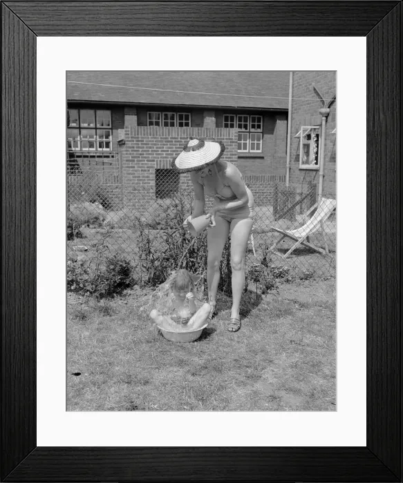 Framed Print : A woman cools her son down by pouring a bucket of cold water over him as he sits out in