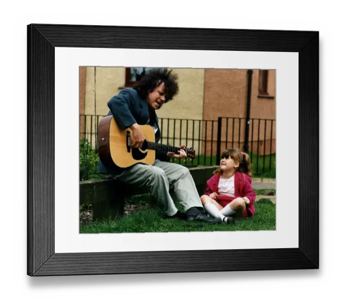 Framed Print : Donovan playing guitar and singing on low wall watched by small girl while on visit to C