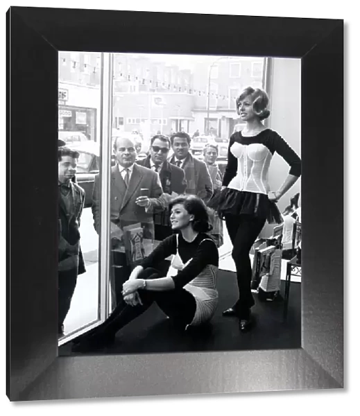 Two women modelling corsets in shop window while people on the street look in - March