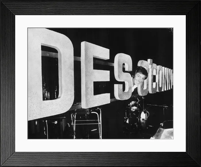 Framed Print : Des O Connor TV Presenter Comedian in fun pose in between enlarged name logo banner