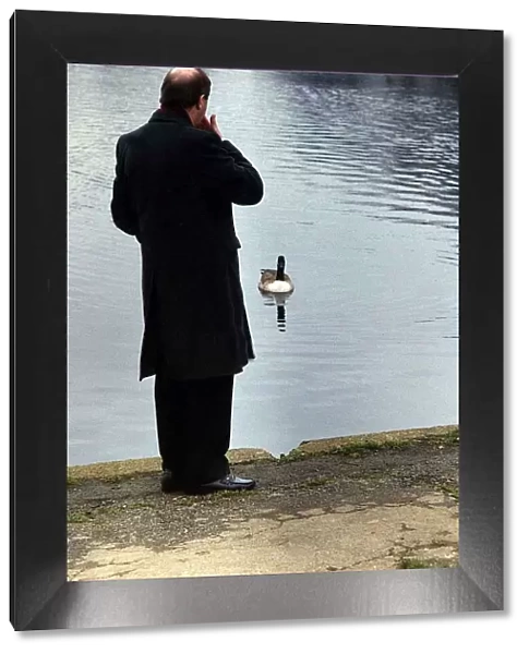 Gorden Kaye actor looking out over a boating lake in a park with duck paddling towards
