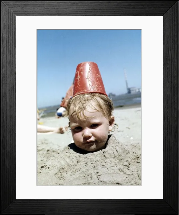 Framed Print : Summer holidays July 1976 - Boy buried in sand with red plastic Bucket on his head