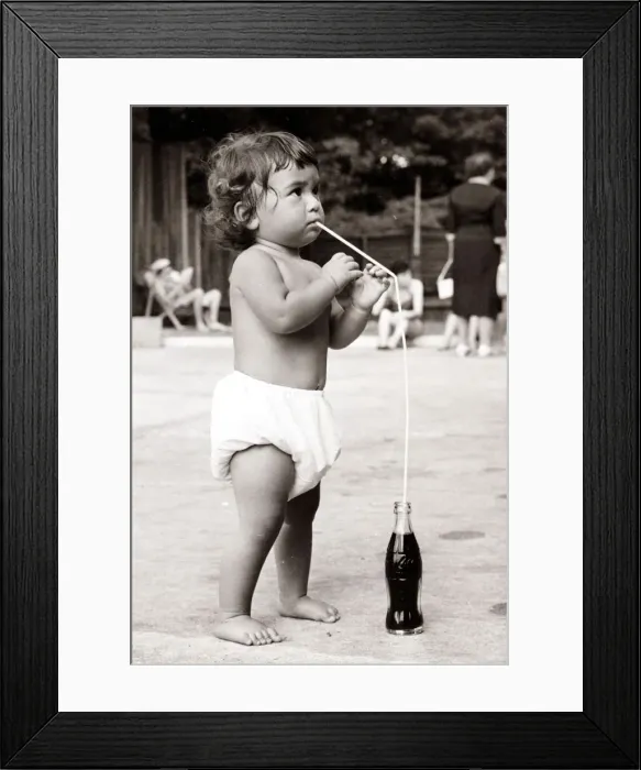 Framed Print : Baby drinking coke with a straw on the beach. Circa 1950
