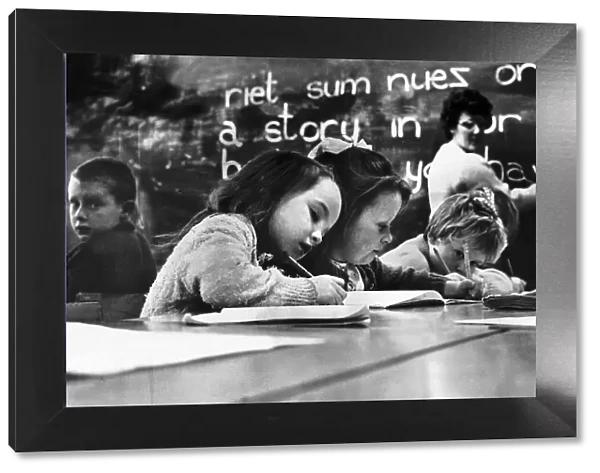 Primary School Children writing at desk with teacher in the background writing on a