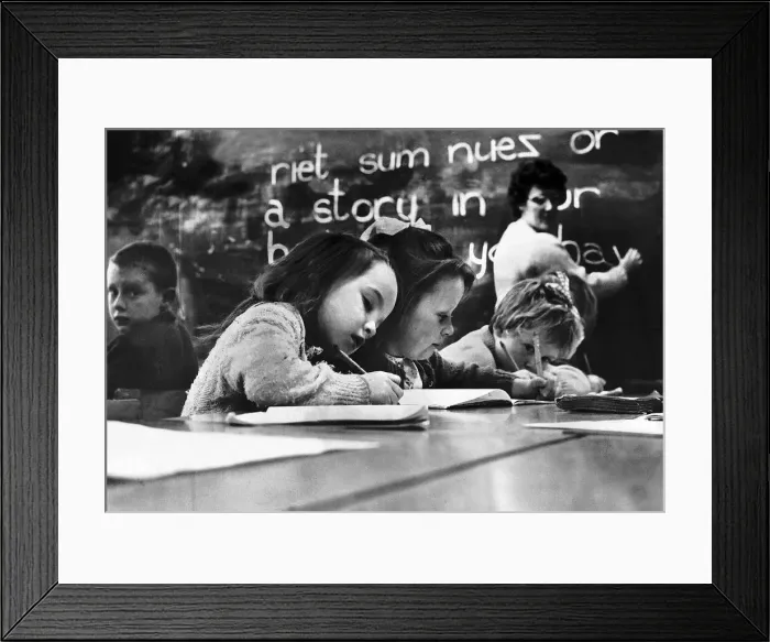 Framed Print : Primary School Children writing at desk with teacher in the background writing on a