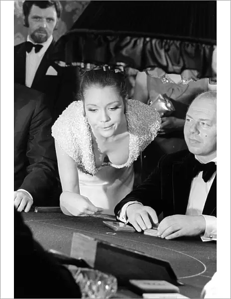 Diana Rigg in a low cut dress leans across the Black Jack table in a casino showing off