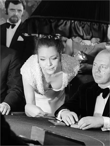 Diana Rigg in a low cut dress leans across the Black Jack table in a casino showing off