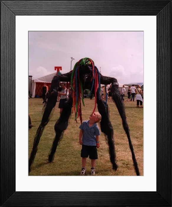 Framed Print : Stilted figure towering over a small boy at T in the Park wearing a bright coloured wig