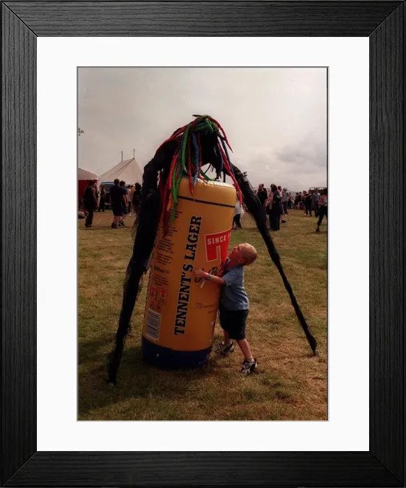 Framed Print : Stilted figure at T in the Park wearing a bright coloured wig standing over an inflatable Framed Print : Stilted figure at T in the Park wearing a bright coloured wig standing over an inflatable