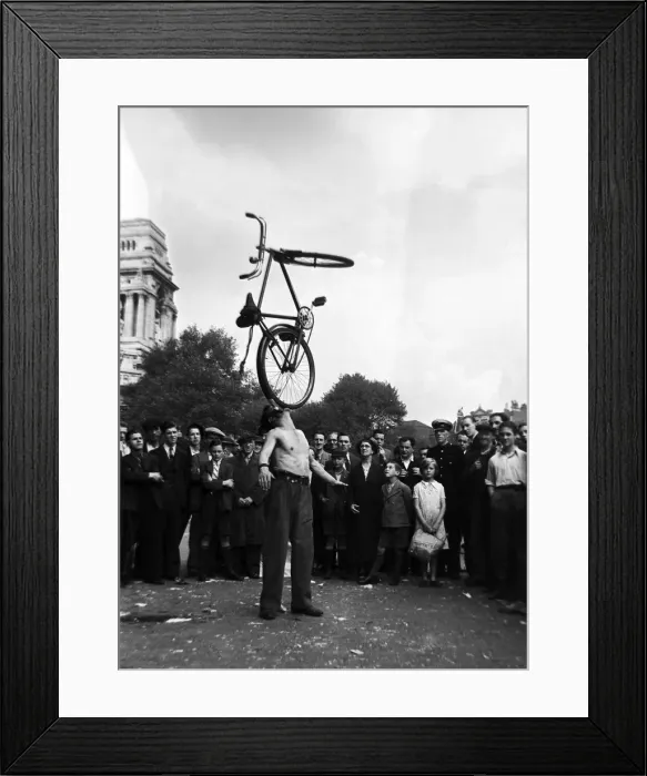 Framed Print : A street entertainer at Tower Hill, London balancing a bicycle using his mouth watched by