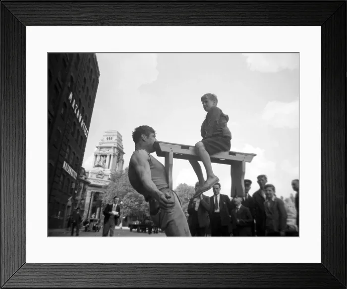 Framed Print : A street entertainer at Tower Hill, London, holding the weight of a boy sitting on a Framed Print : A street entertainer at Tower Hill, London, holding the weight of a boy sitting on a