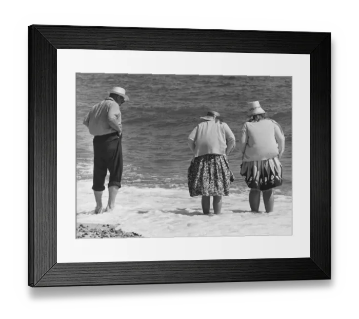 Framed Print : Two old women and one old man with hats stand on the beach with their feet in the water