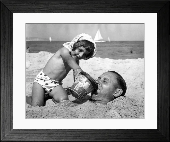 Framed Print : 4 year old Gloria Smart gives her dad a drink of seawater after burying him in the sand Framed Print : 4 year old Gloria Smart gives her dad a drink of seawater after burying him in the sand