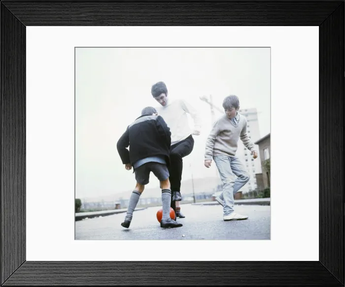 Framed Print : Manchester United footballer George Best playing football in the street with two young