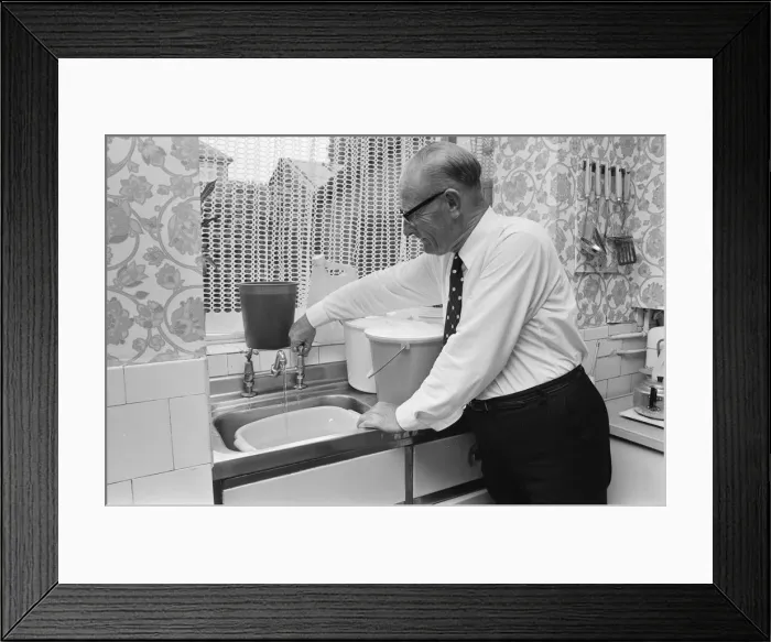 Framed Print : An elderly man running the tap in his kitchen, filling up buckets as a water shortage