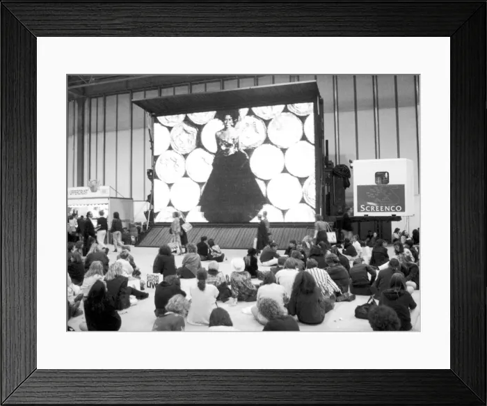 Framed Print : Clothes Show Live, visitors to the clothes show relax beneath one of the giant video