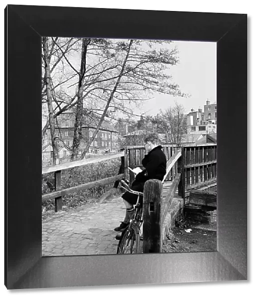 A boy reading a book by a bridge crossing the River Wey, in Guildford, Surrey. April 1954
