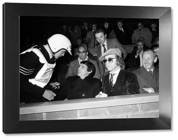 A policeman talking to Graham Taylor and Elton John, who are watching the football match