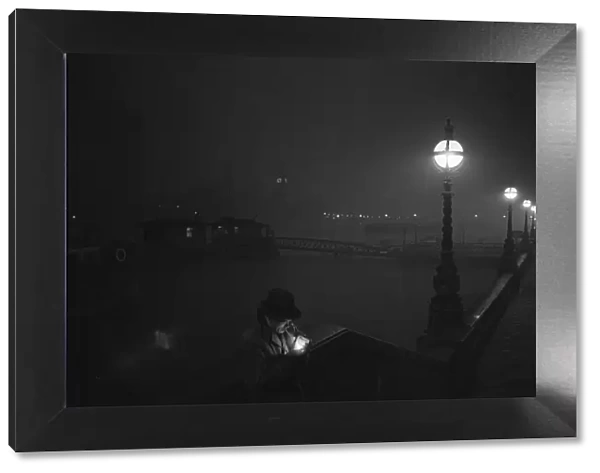 A man lights a cigarette on the Thames Embankment at night. Circa 1955