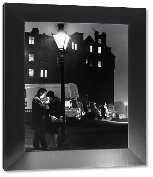 A man lighting a cigarette for a woman, standing under a street lamp at night