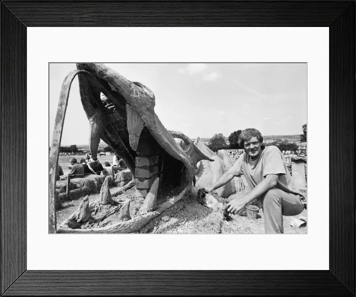 Framed Print : Glastonbury Festival, Pilton, Somerset. A man sits with a wooden model of a Framed Print : Glastonbury Festival, Pilton, Somerset. A man sits with a wooden model of a