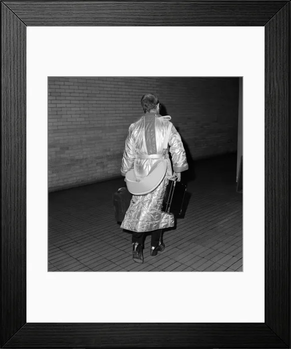 Framed Print : Rock star Elton John at Heathrow Airport. He is wearing a long silver nylon quilted