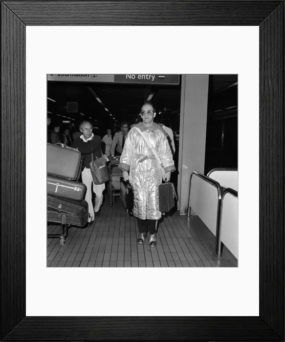 Framed Print : Rock star Elton John at Heathrow Airport. He is wearing a long silver nylon quilted Framed Print : Rock star Elton John at Heathrow Airport. He is wearing a long silver nylon quilted