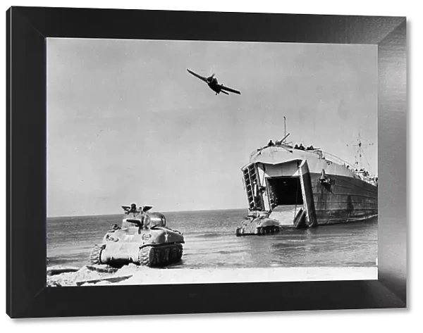 A Wildcat plane passes over a US Navy Landing Ship as it discharges a Sherman tank onto a