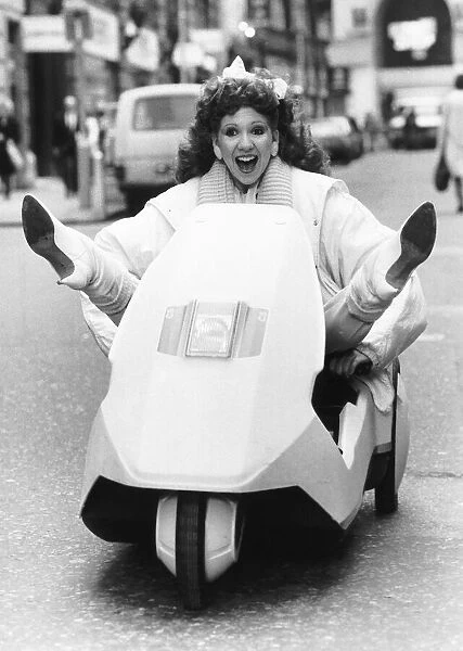 Bonnie Langford sitting in a Sinclair C5 on the road with her feet out up in a v-shape