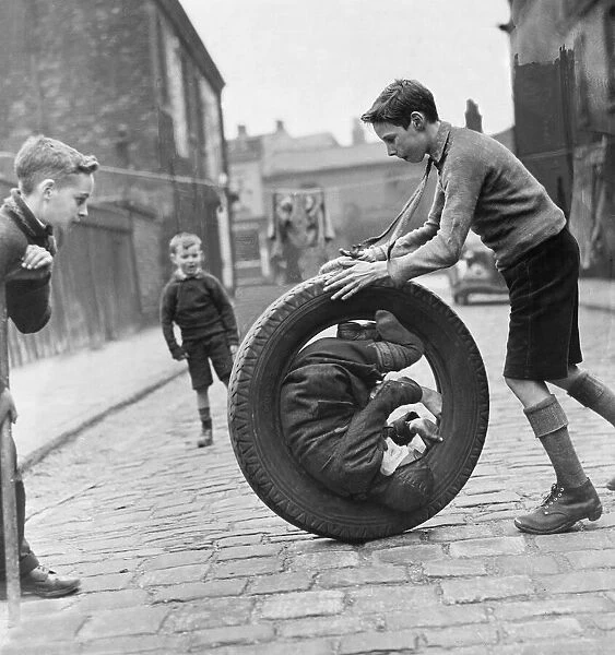 The boys are playing with a car tire, one boy is sitting inside the tire while another