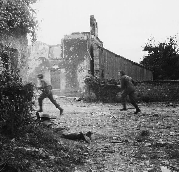 A Bren gunner covers the roadway while men move along the street of the village of Hottot