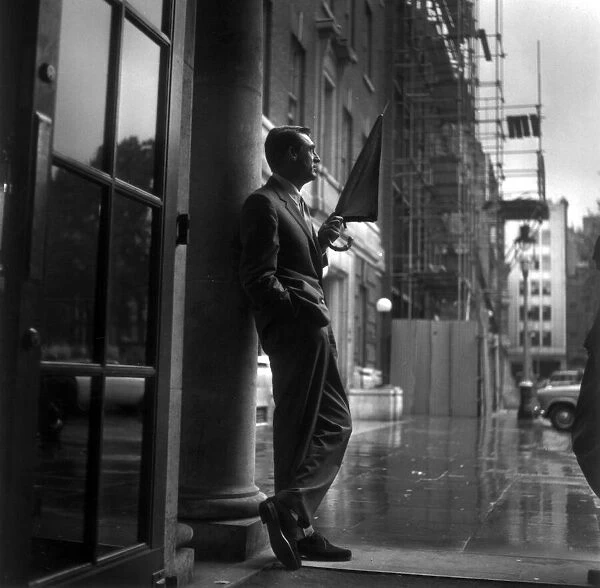 CARY GRANT SHELTERS FROM THE RAIN ON THE PORCH OF A LONDON HOTEL. 1957