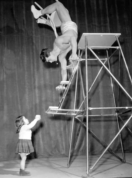 Framed Print : Children Acrobat. A young girl holding a cup looks up at a man walking down steps on his