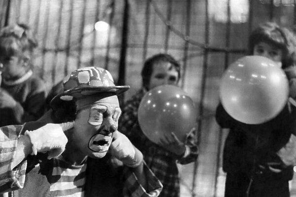 A circus clown puts his fingers in his ears as members of the audience blow up balloons