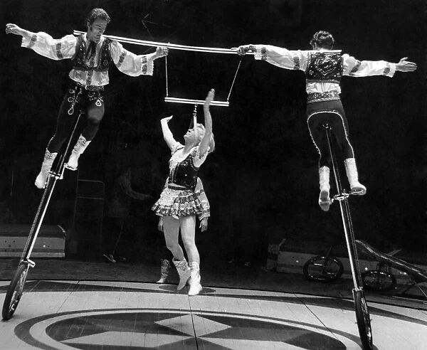 Circus performers on unicycles lifting a woman up between them as they ride around her