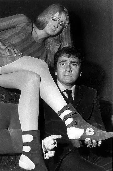 Comedian Dudley Moore pulling a face inspects a pair of boots called 'Sex Puss'